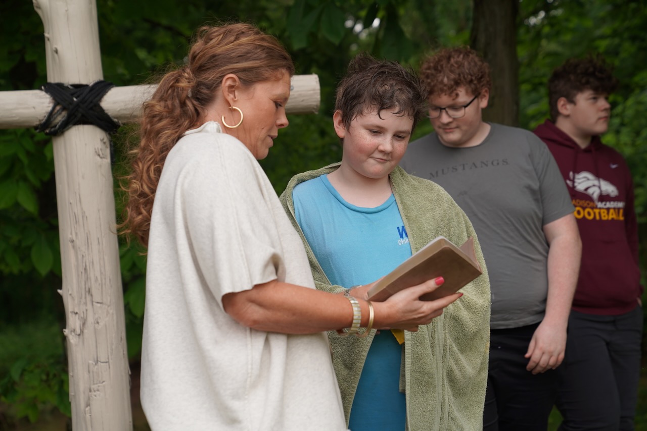 A teacher reads from a the Bible to a young boy wrapped in a towel after being baptized, standing next to a wooden cross outdoors.