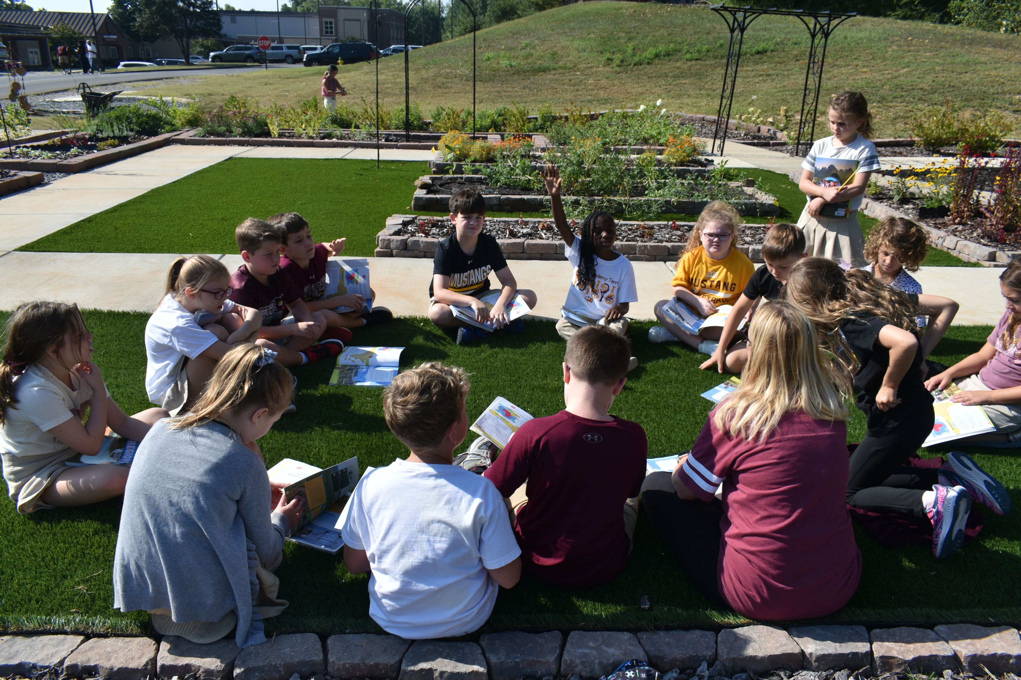 A group of children sit in a circle on a lawn with books for an outdoor class in a garden.