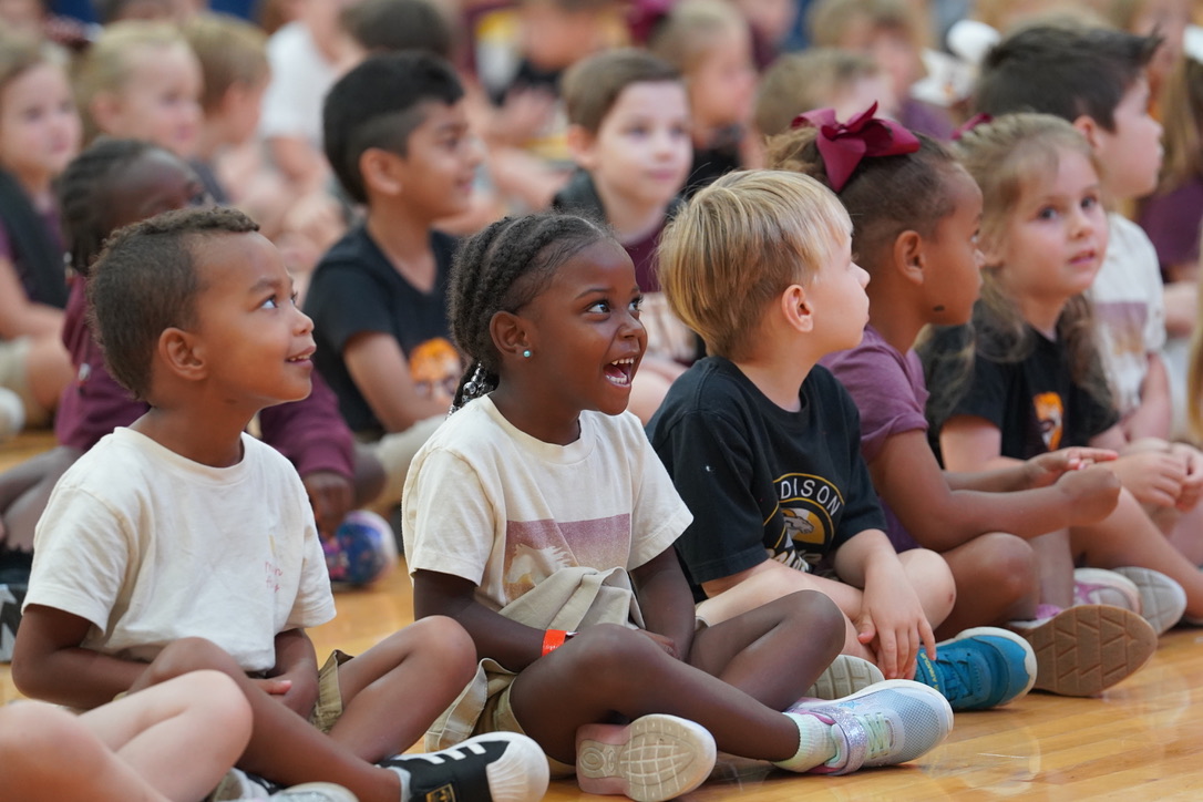 A group of diverse elementary students sits cross-legged on a gymnasium floor, looking on with captivated and happy expressions.