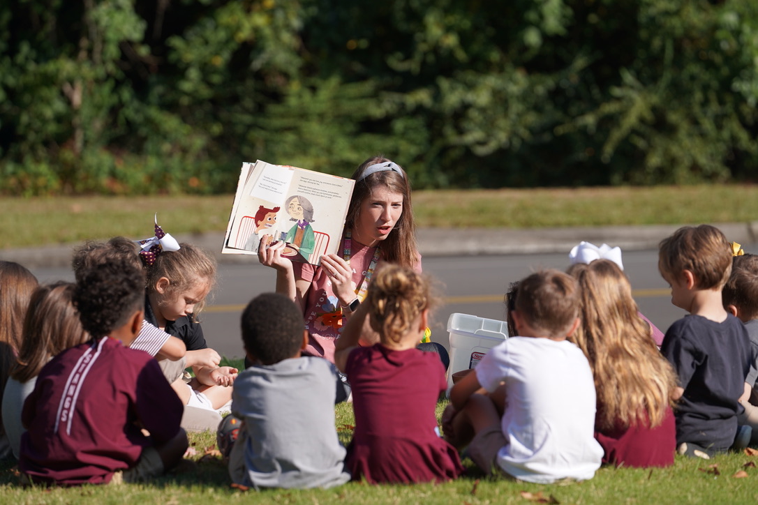 A teacher reads a storybook to a group of young children sitting on the grass outdoors.