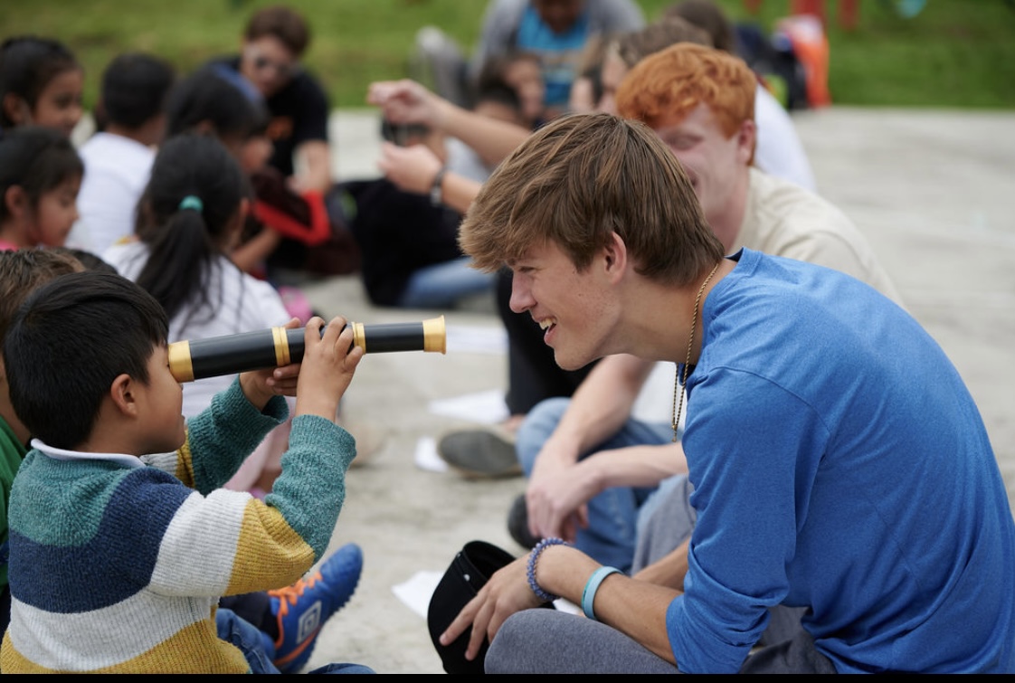 A young boy in a striped sweater looks through a toy spyglass at a smiling teenage boy sitting across from him.