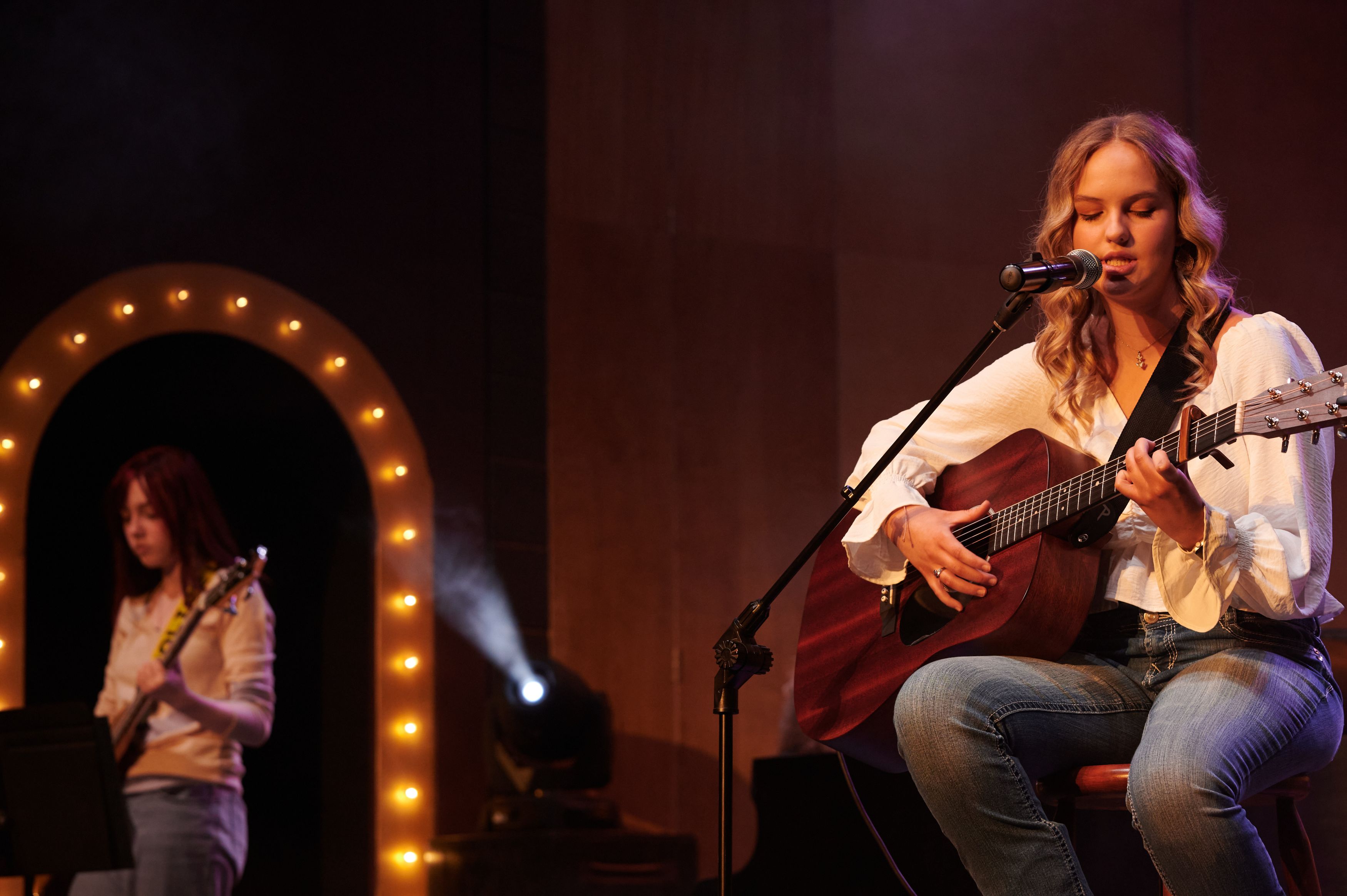 A young woman sings and plays acoustic guitar into a microphone while performing on a dimly lit stage, with a bass guitarist in the background.